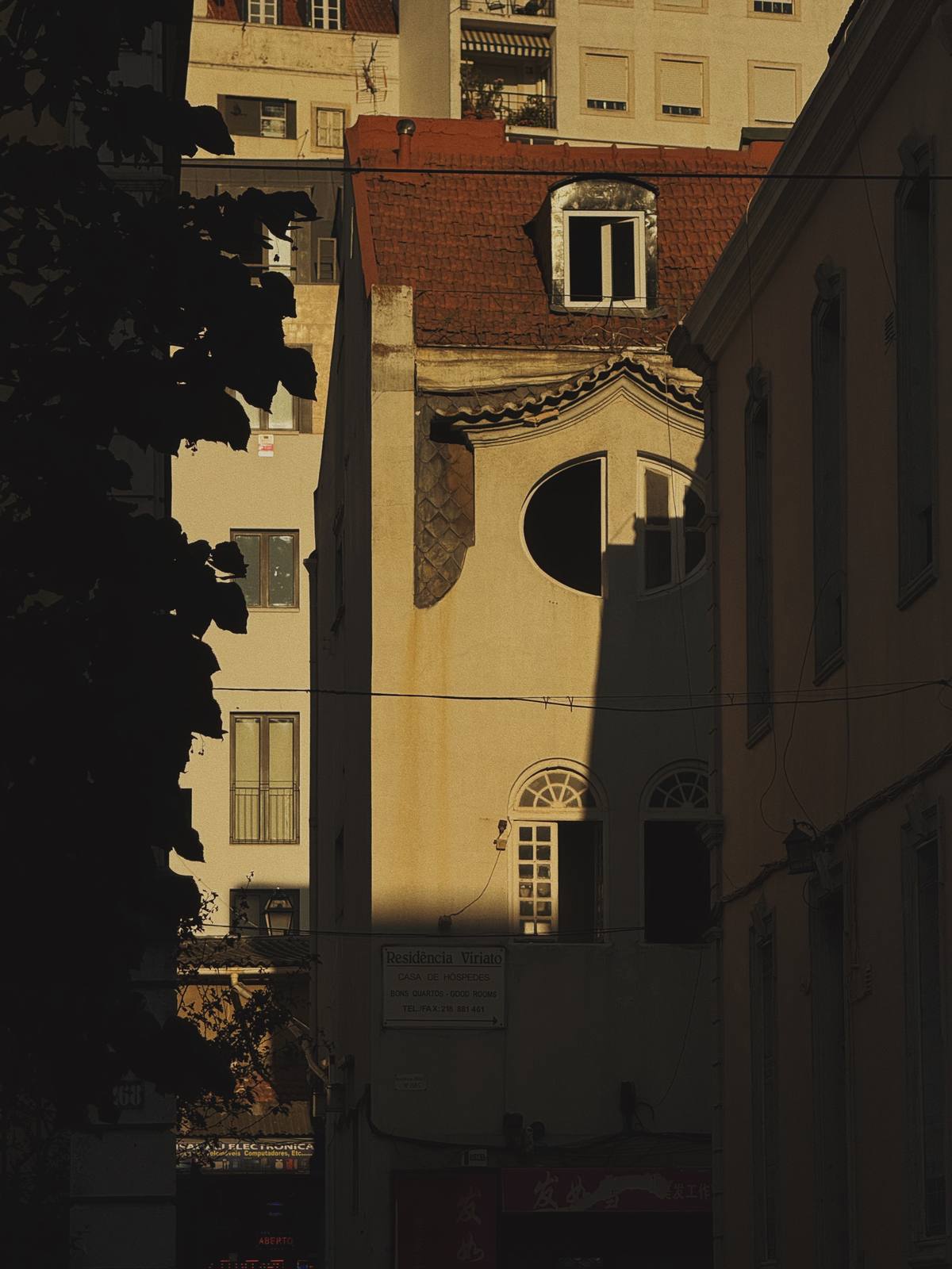 Chapel in Deep Shadow, Graça & Miradouros, Lisbon — Ochre chapel facade with an oval eye window caught in a shaft of late light, a dark silhouetted tree framing — Lisbon seen in architectural layers, 