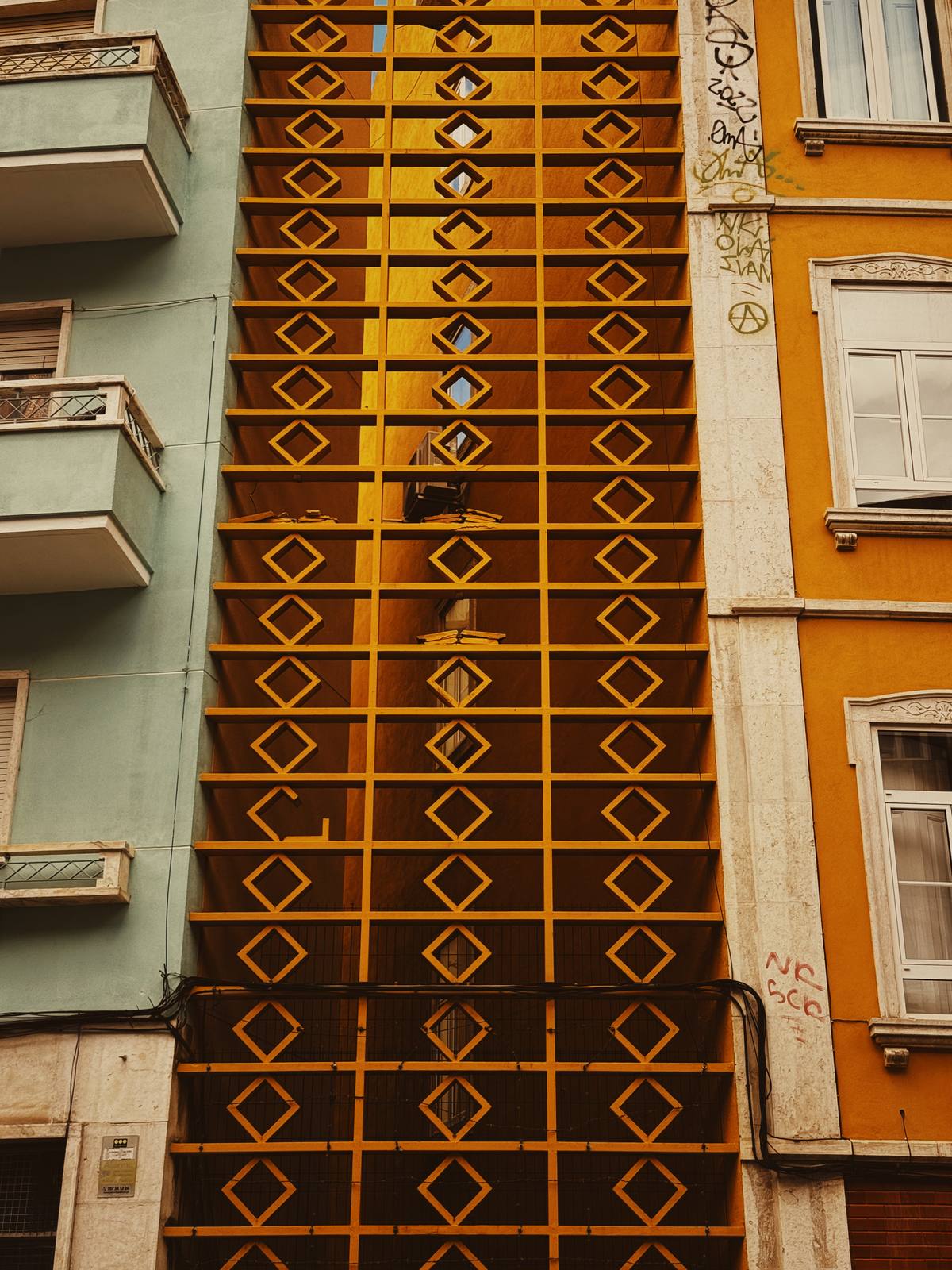 Diamond Screen, Yellow Wall, Graça & Miradouros, Lisbon — A graphic diamond-lattice facade in deep gold fills the frame between mint and orange flanking walls — bold pattern, brand-yellow palette.