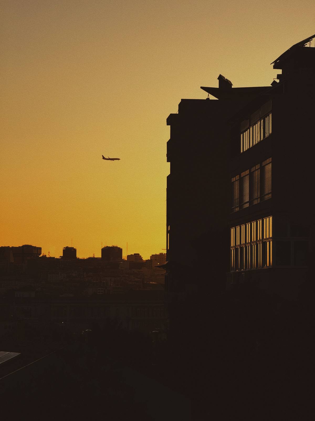 Plane Over Yellow City, Graça & Miradouros, Lisbon — A tiny aircraft suspended in a pure yellow sunset sky beside a dark building silhouette — the plane reads as an isolated figure against vast colour