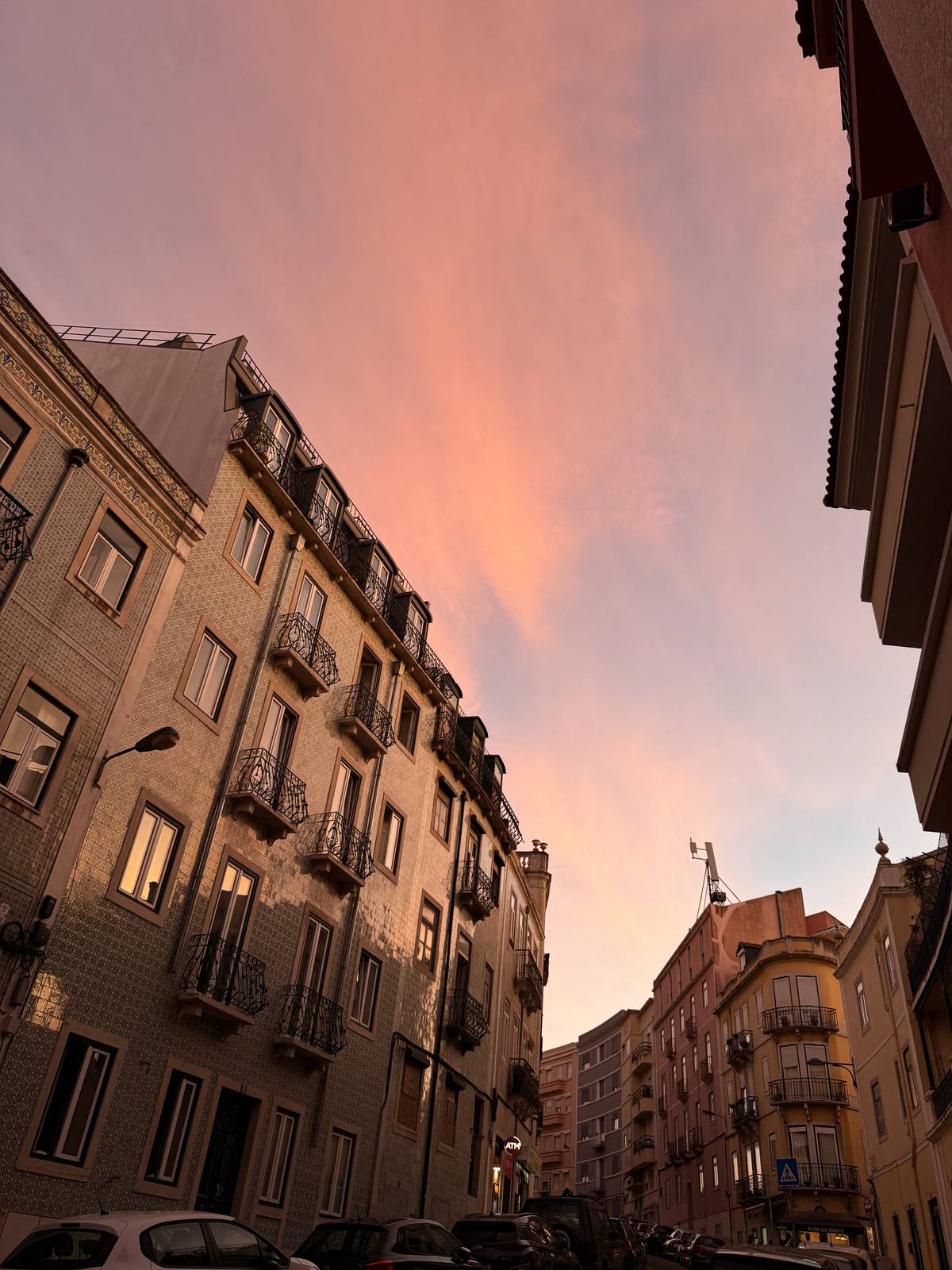 Azulejos Into Pink Sky, Graça & Miradouros, Lisbon — Upward worm's-eye on a tiled facade with wrought-iron balconies, rose-pink dusk filling the void — geometry and colour working together.