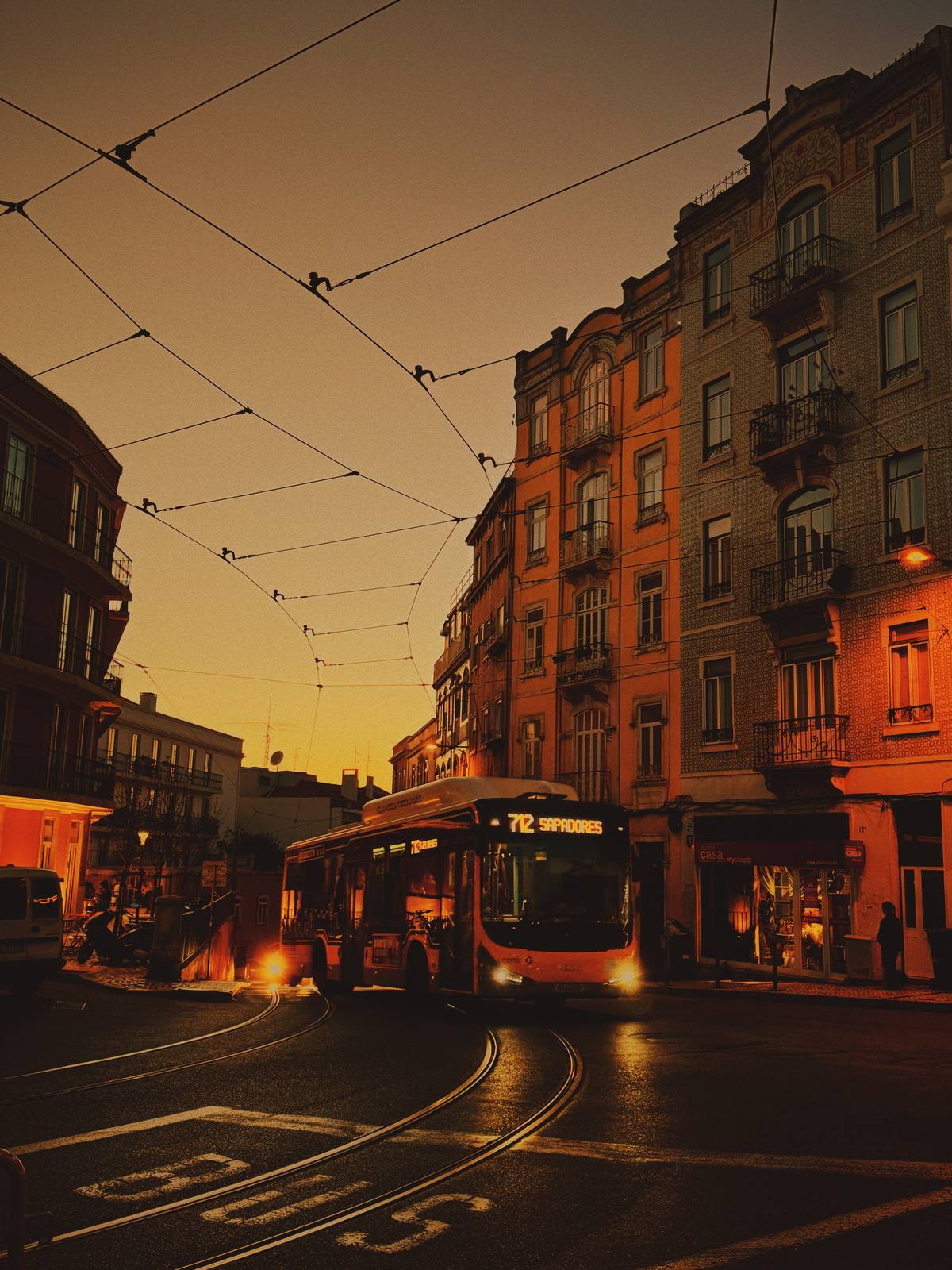 Bus 712, Burning Gold, Graça & Miradouros, Lisbon — A bus navigates the junction at deep golden hour — overhead wires, amber-lit ornate facade, tram rails wet on the road; infrastructure as pure atmos
