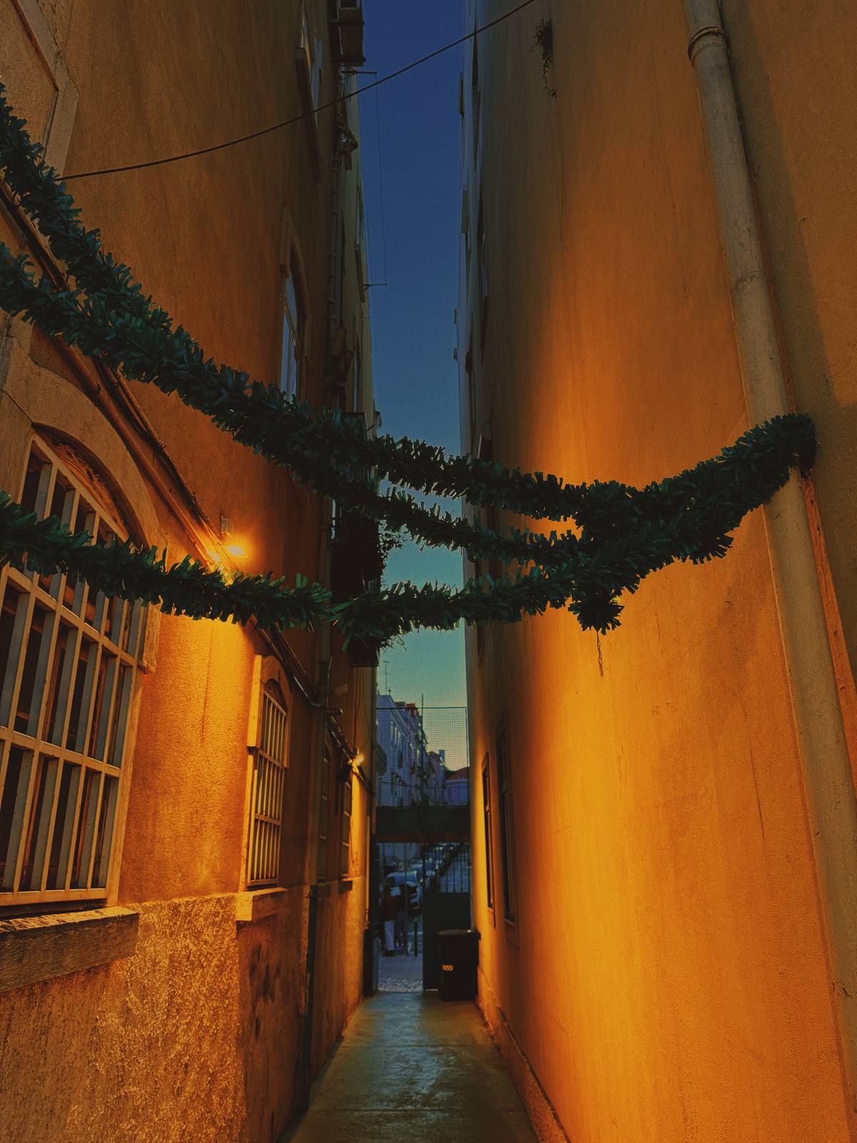 Garland Alley, Blue Hour, Graça & Miradouros, Lisbon — Narrow passage with warm orange walls and a garland strung across, leading the eye to a sliver of blue-hour street beyond — warm-cold colour tens