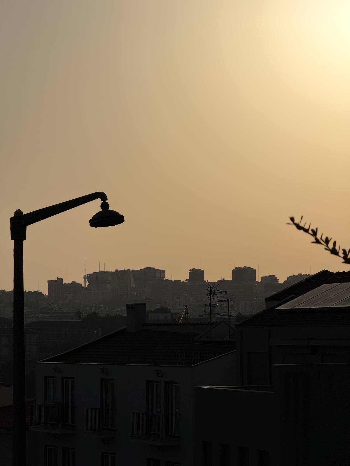 Lamp Over the Haze, Graça & Miradouros, Lisbon — Cast-iron street lamp silhouetted against amber sunset haze with the Lisbon skyline receding in three tonal layers — bold graphic anchor, contemplative