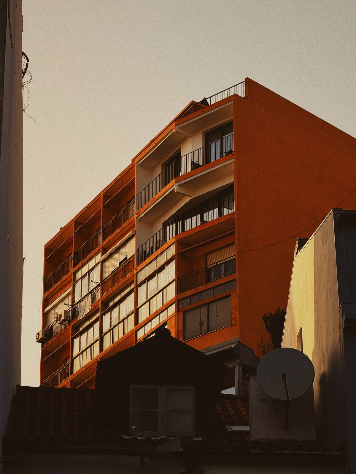 Orange Block, Pale Sky, Graça & Miradouros, Lisbon — Deep terracotta residential slab glowing in late sun against a near-white sky — bold graphic shape, strong tonal contrast, the satellite dish groun