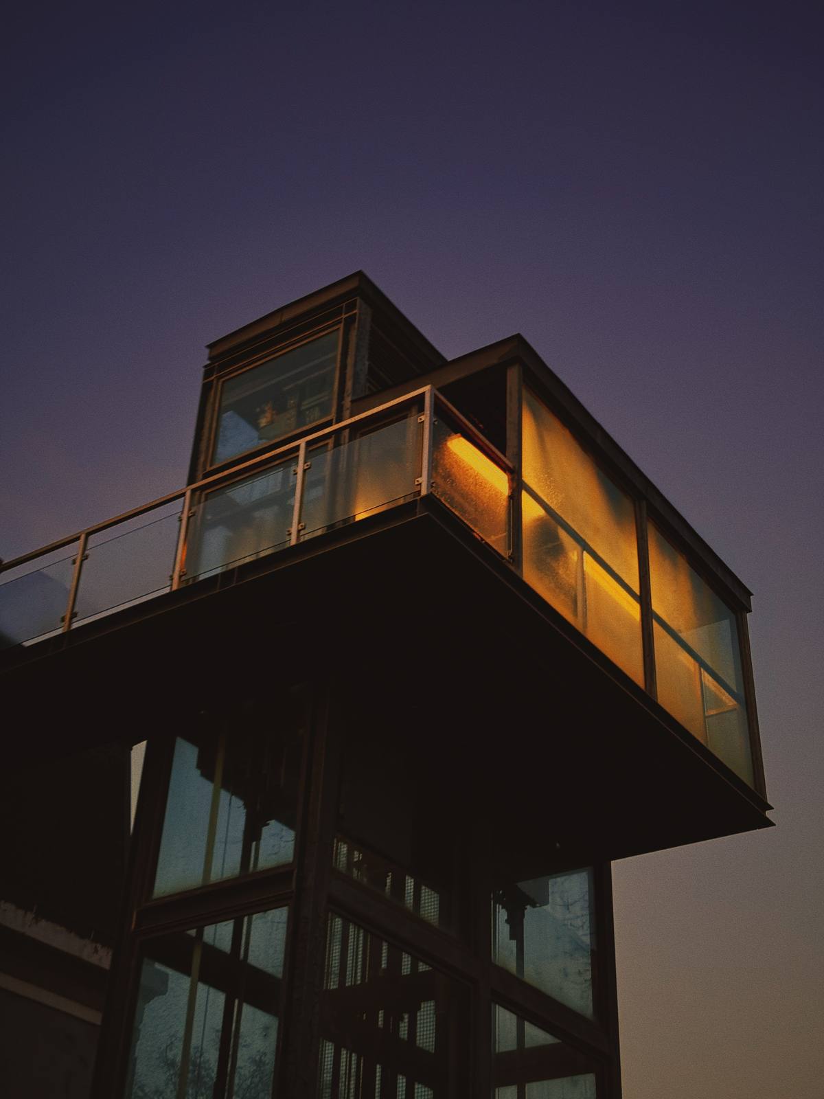 Amber Box, Violet Sky, Cais do Sodré, Lisbon — Glass viewing platform glows amber from within against a deep purple dusk — isolated architecture, quiet luminosity, perfect tonal restraint.