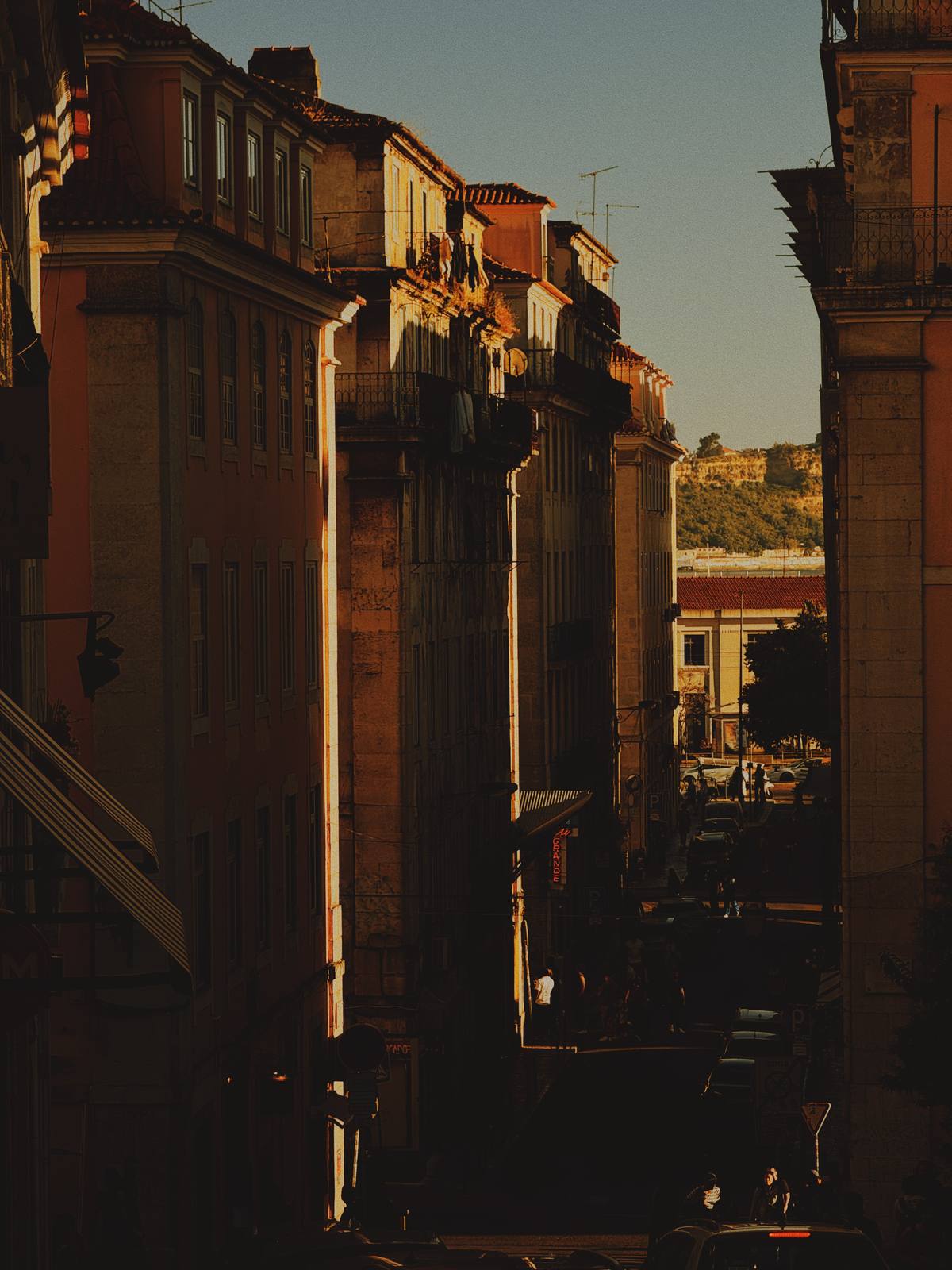 The Hill Behind the Street, Cais do Sodré, Lisbon — Compressed telephoto down a descending rua at golden hour — buildings in amber shadow, castle hill visible in the sliver of light at the end, Lisbon