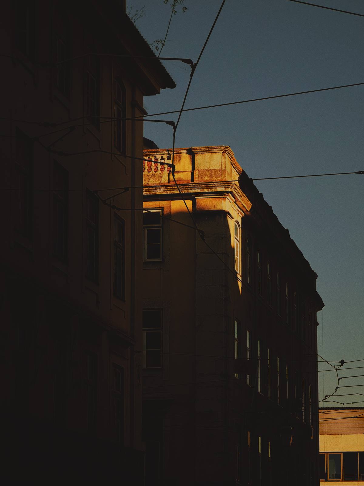 One Window, Last Sun, Cais do Sodré, Lisbon — A single golden cornice fragment glows between deep shadow masses and tram wires — bold chiaroscuro with strong Lisbon atmosphere.