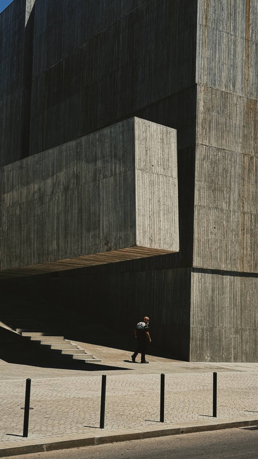 Small Figure, Vast Concrete, Cais do Sodré, Lisbon — Single figure beneath a cantilevered brutalist block — shadow geometry is immaculate, the human scale against the mass is the brand's core tension.