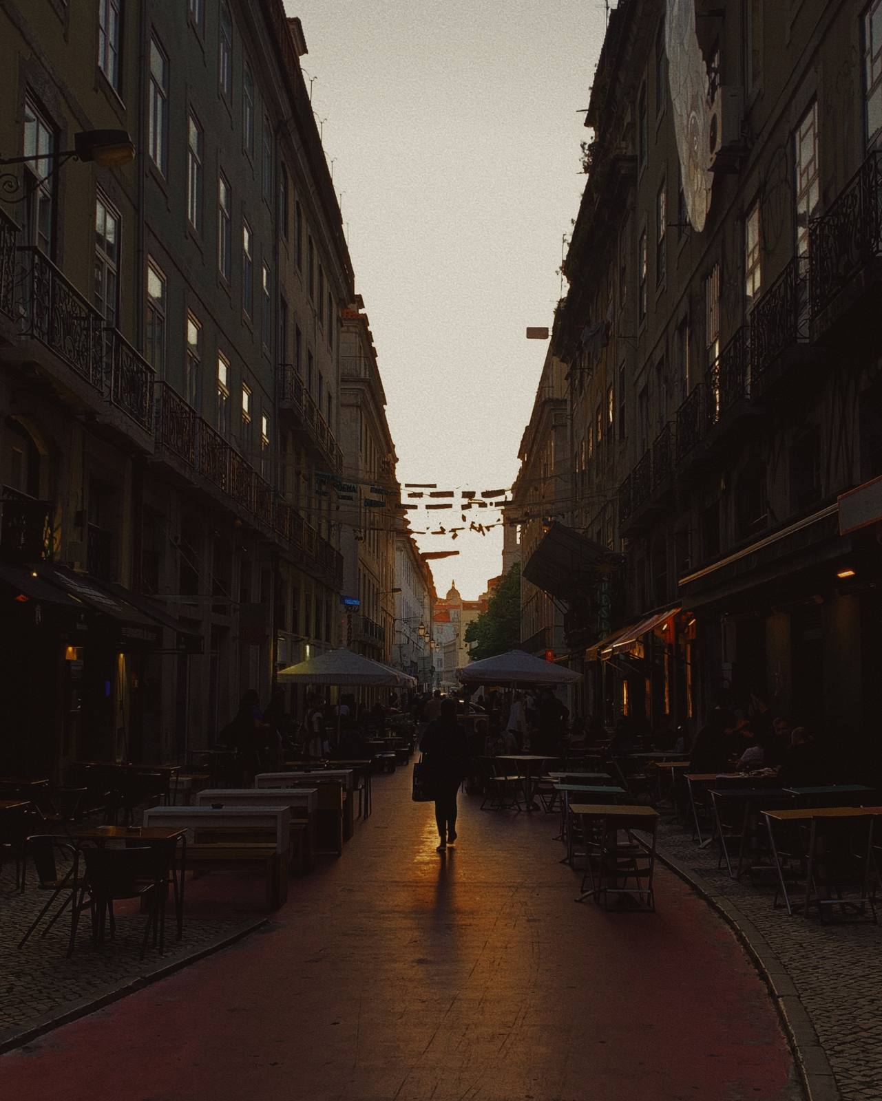 One Figure, Pink Street, Cais do Sodré, Lisbon — Perfect canyon perspective down Rua Nova do Carvalho at golden hour — lone silhouette walking toward burning light, symmetric flanking buildings, stron
