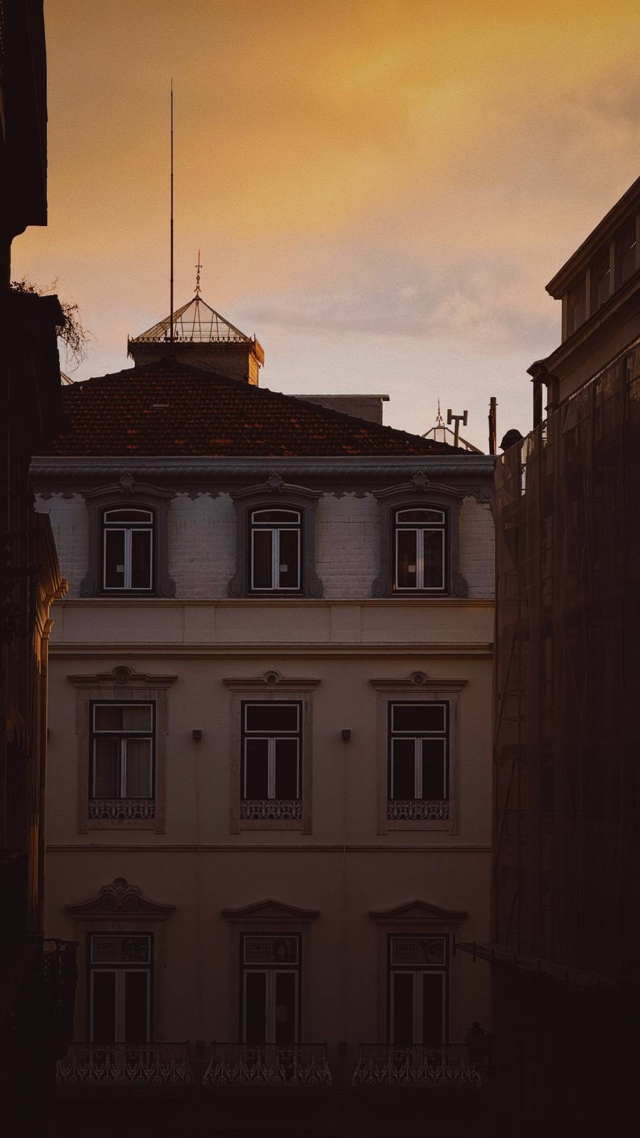 Golden Sky Over the Emenda, Chiado, Lisbon — Belle-époque facade pinched between dark flanking buildings, golden sunset sky — Lisbon in layers, pure ABIASSI palette.