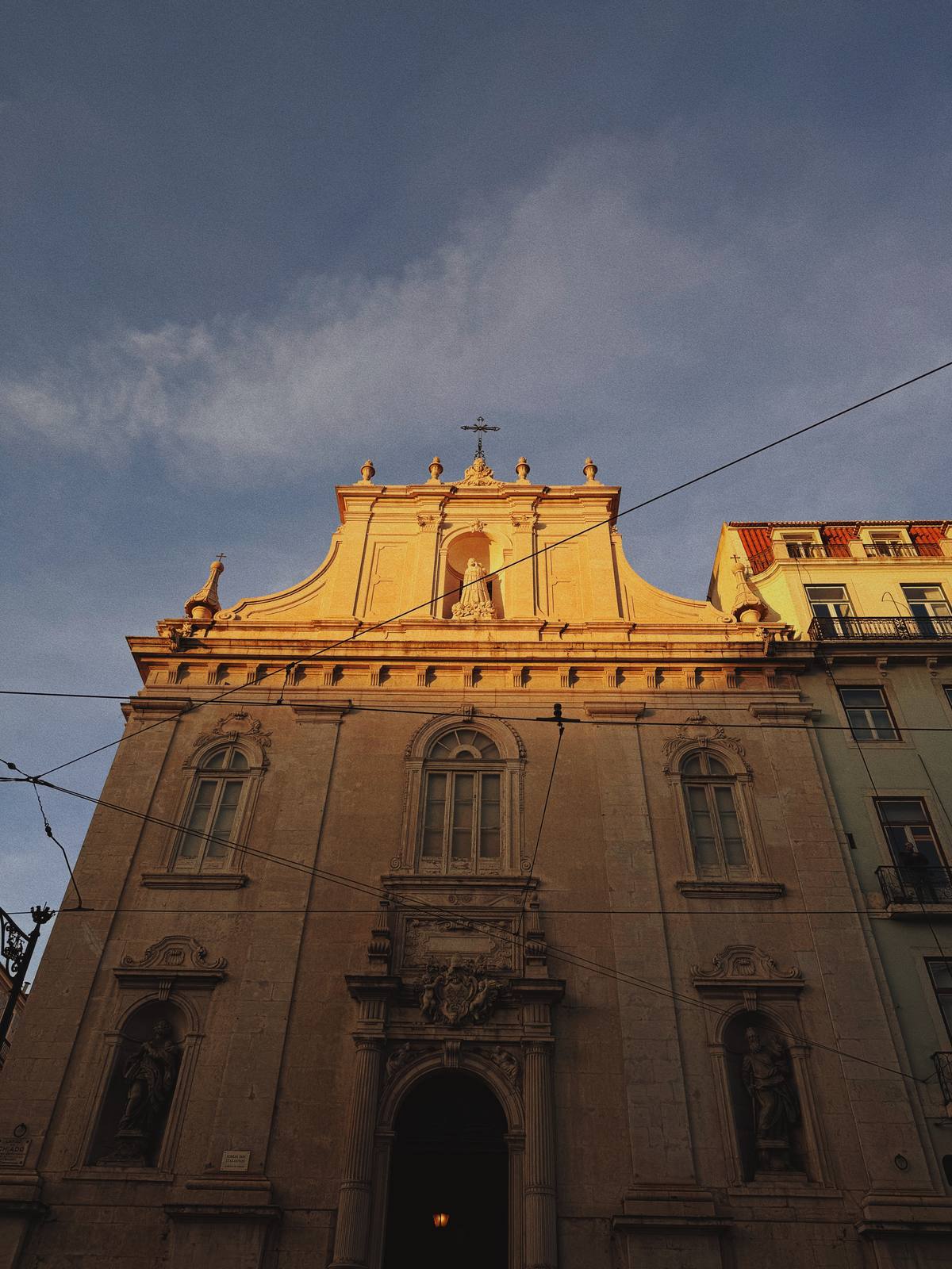 Mártires in Last Light, Chiado, Lisbon — Belle-époque church facade in raking sunset gold against blue-grey cloud sky; bold upward composition, warm stone against cold sky, no distractions.