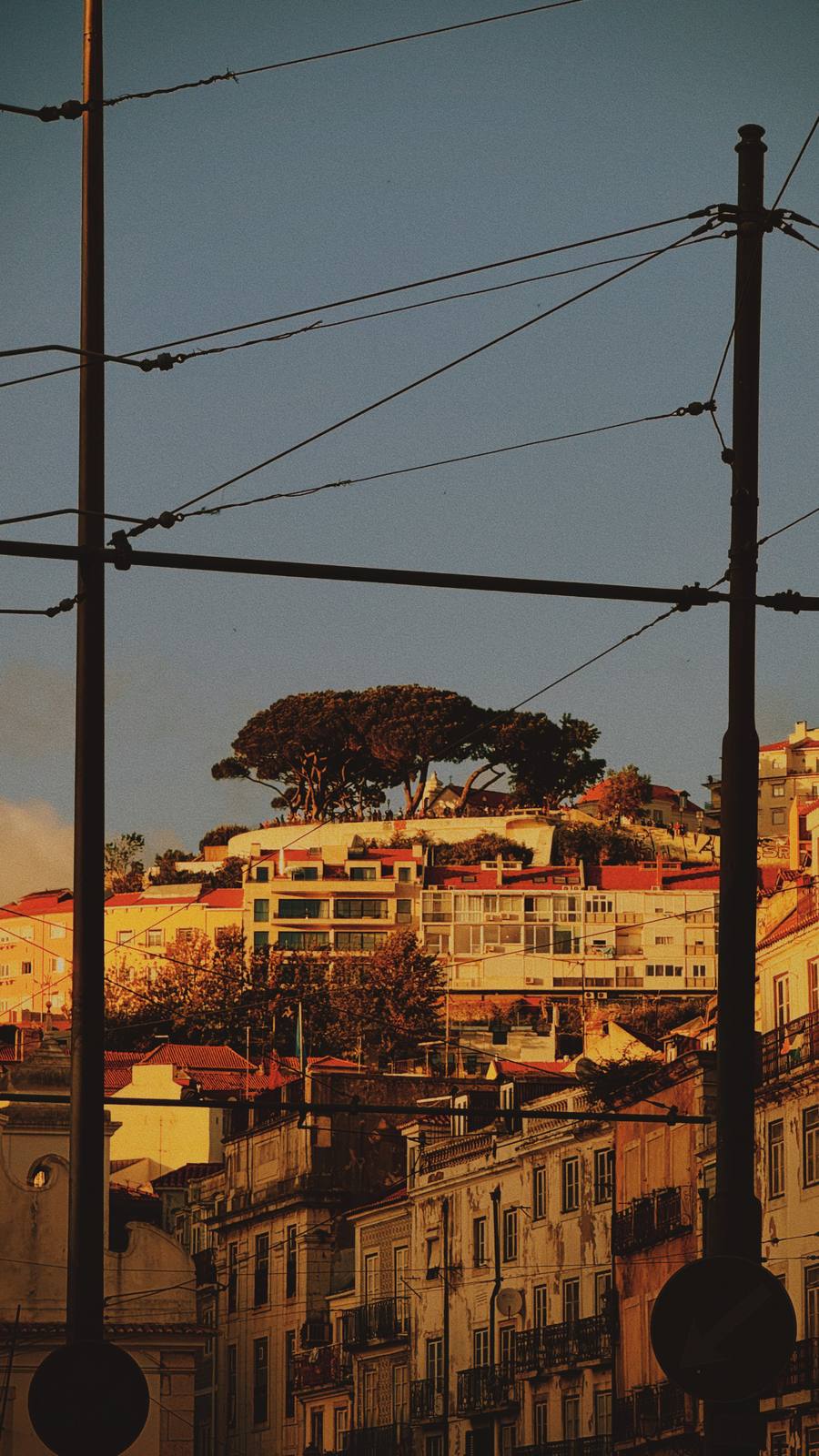 Wires Over the Hill, Mouraria, Lisbon — Tram poles and crossing wires layer the frame; warm-lit hillside city rises behind in the golden hour — Lisbon seen in strata.
