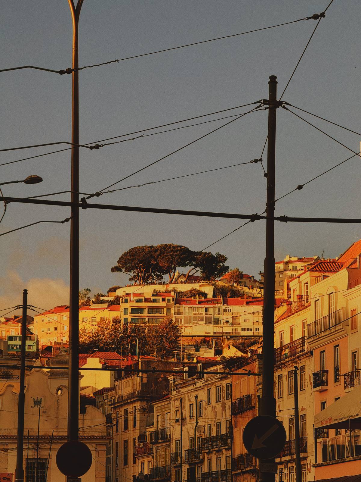 Cables Over Golden Lisbon, Mouraria, Lisbon — Tram cables form deliberate graphic geometry over a warm amber hillscape at golden hour — layered city depth with strong vertical poles as anchors.