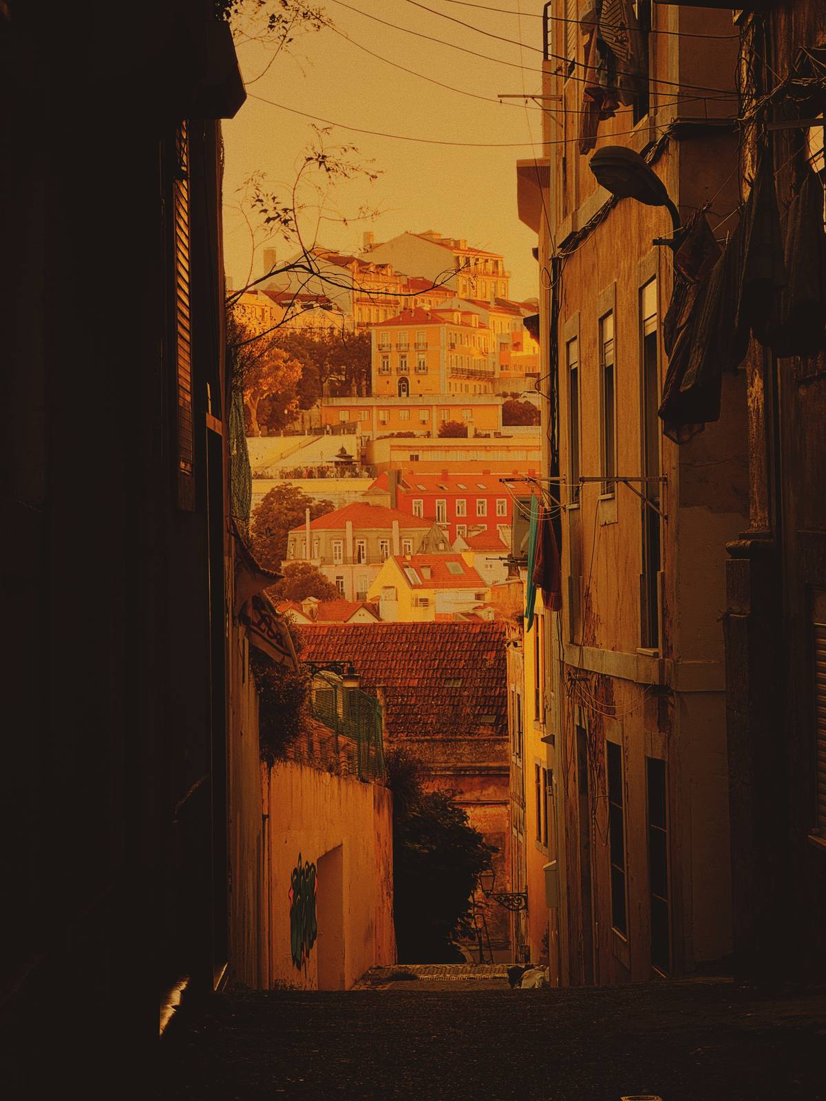 Amber Lane, City Beyond, Mouraria, Lisbon — Steep alley frames Lisbon rooftops in deep amber light — classic Mouraria layering, strong silhouette walls, no clutter.
