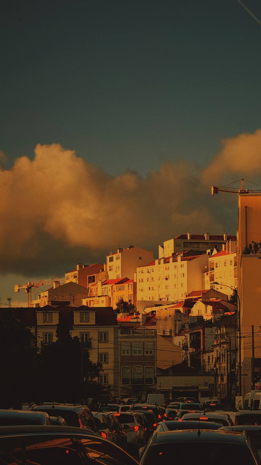 Hill Rising, Storm Above, Mouraria, Lisbon — Martim Moniz at golden hour: city terraces rising in layers to the hill, dramatic storm clouds catching last amber light, tram wires and cranes as vertical