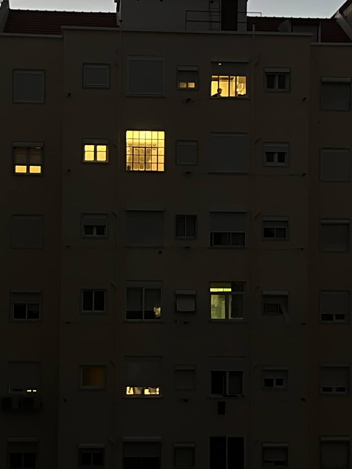Neighbours After Dark, Penha de França, Lisbon — A night facade of staggered warm windows, a faint figure in one — quiet surveillance of ordinary private life.