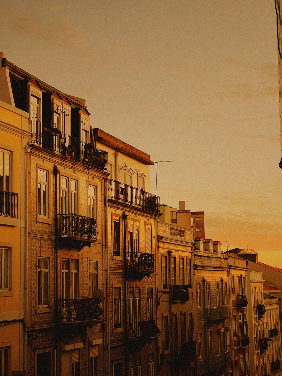 Balconies Into Amber Dusk, Penha de França, Lisbon — Lisbon street facade with iron balconies receding into a deep amber sunset — colour and perspective both portfolio-grade.