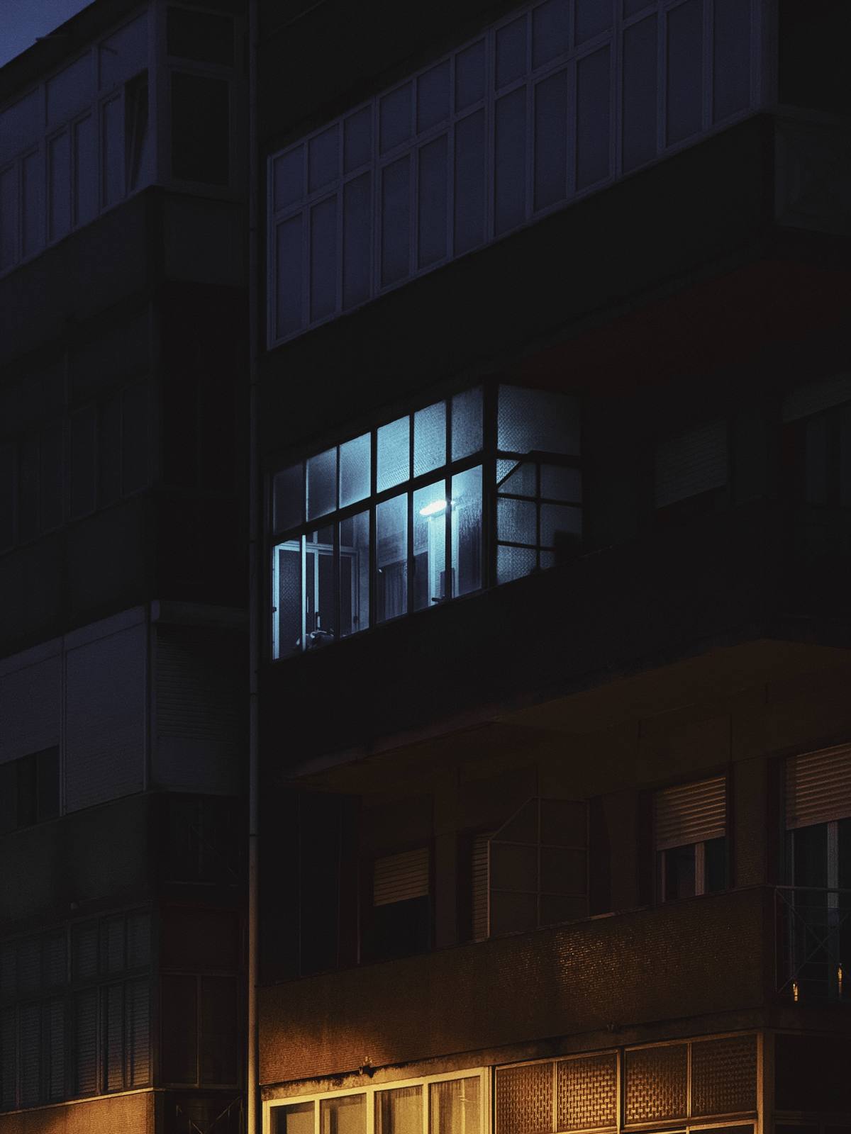 One Light, Everyone Asleep, Penha de França, Lisbon — A single blue-lit window glowing in a near-black facade at night — quiet narrative tension, strong isolation.