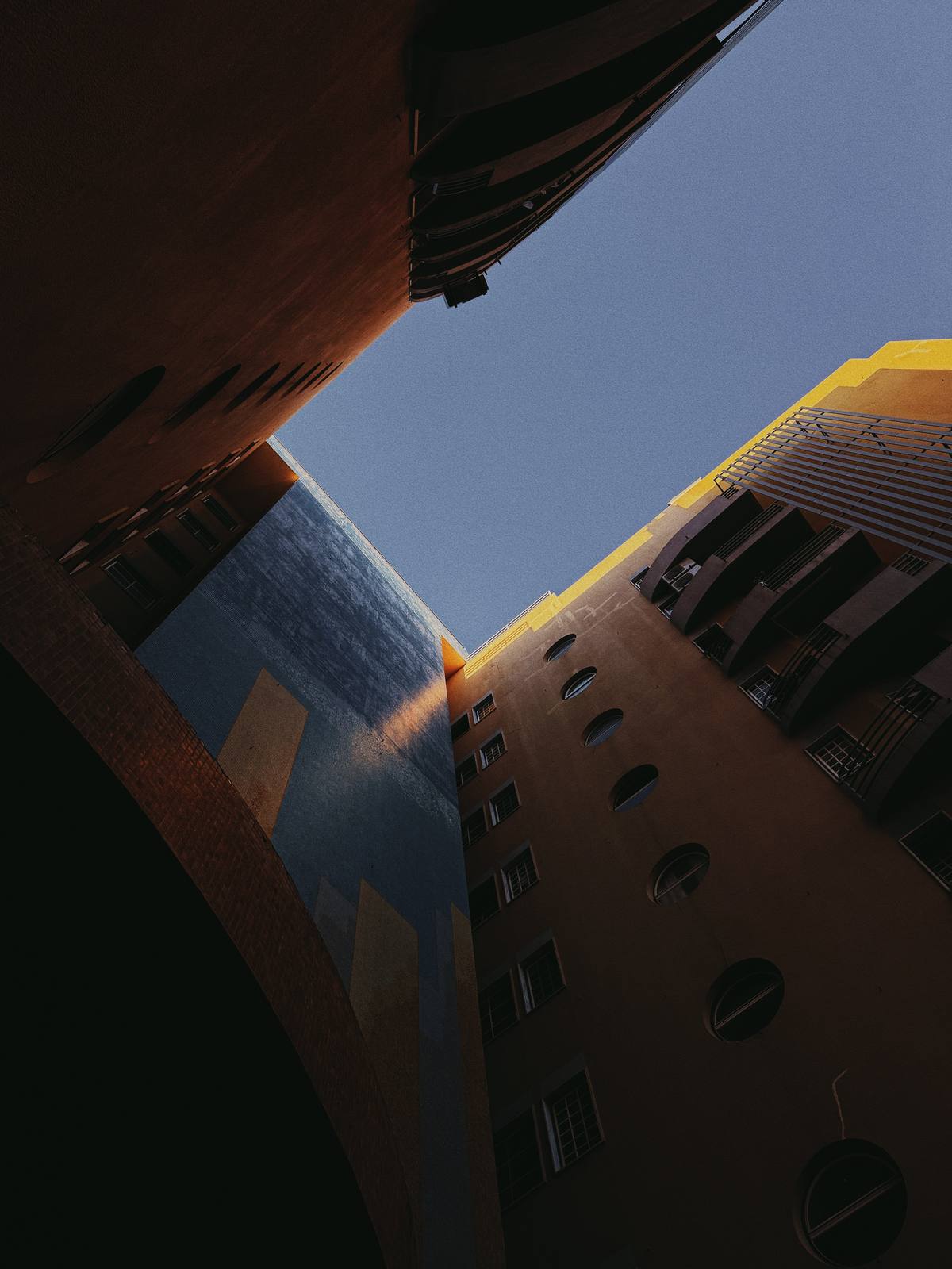 Courtyard, Open Sky, Penha de França, Lisbon — Worm's-eye view between three buildings catches a yellow wall and round portholes — strong geometry, companion to the triangle frame.