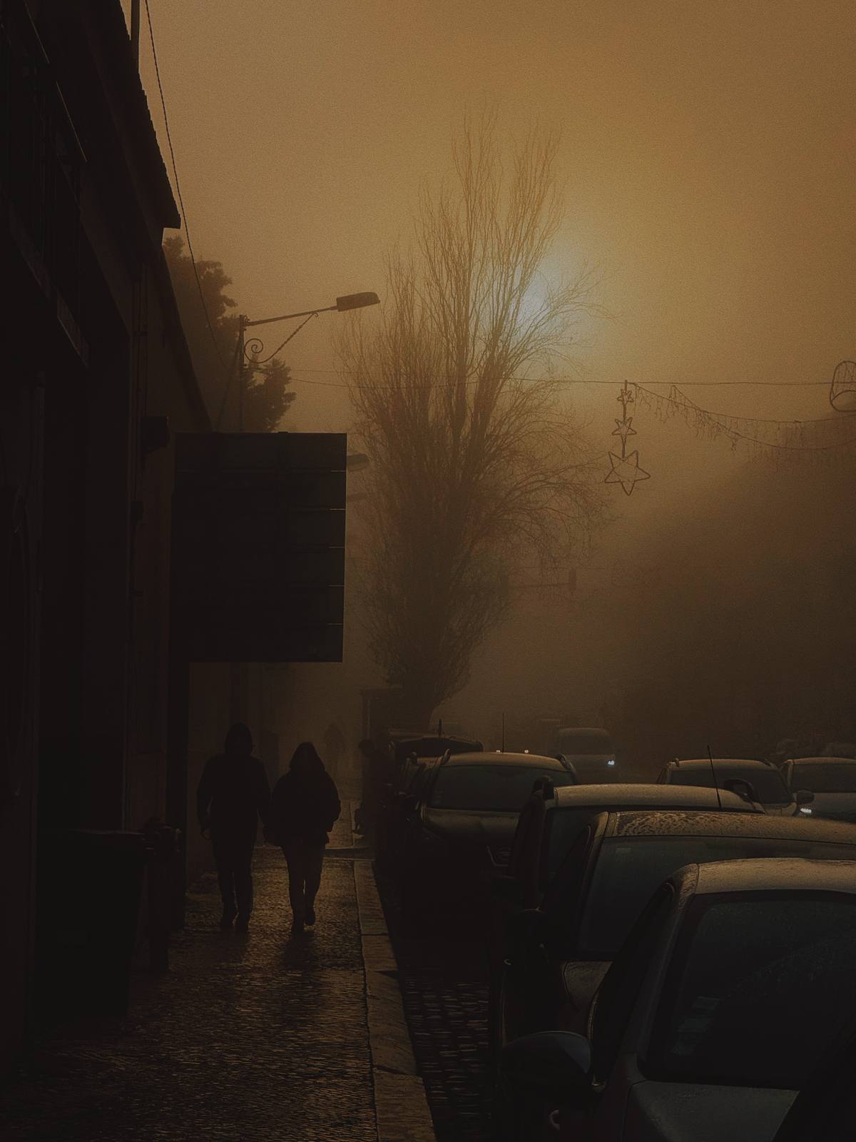 Two Figures, Amber Fog, Penha de França, Lisbon — Dense winter fog turns the street amber; two silhouettes recede — the isolated human form in architectural space, the brand thesis exactly.